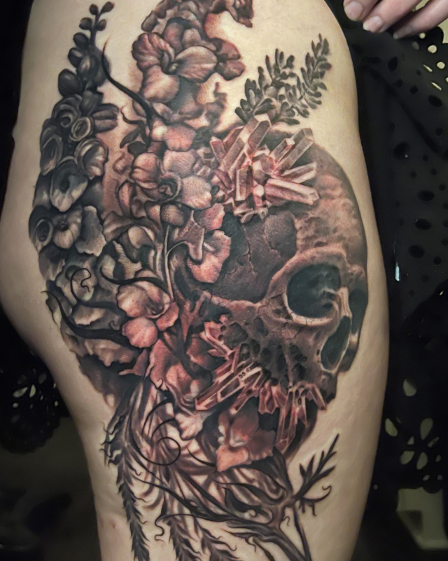 Skull with Flowers and crystals erupting from it thigh tattoo done in black and grey fine line ink by Shaine Smith of Sacred Mandala Studio.